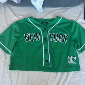 Grön baseballtröja från FB Sister - Säljer en cool grön baseballtröja från FB Sister med texten 'New York' och siffran '92' på framsidan. Tröjan har korta ärmar och knappar framtill. Perfekt för en sportig look!