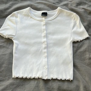 Vit ribbad kortärmad topp från Gina Tricot - Säljer en söt vit ribbad topp från Gina Tricot i storlek XS. Toppen har korta ärmar och knappar framtill, med en fin vågig kant vid ärmslut och nederkant. Perfekt för en stilren look!