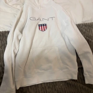 Vit hoodie från GANT - Säljer en stilren vit hoodie från GANT med broderad logga och emblem på bröstet. Tröjan har en praktisk magficka och långa ärmar. Perfekt för en avslappnad stil.  Använd en eller två gånger. Pris kan även diskuteras 