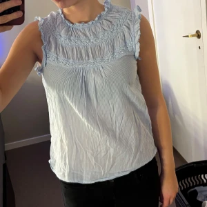 Ljusblå blus med spetsdetaljer - Säljer en söt ljusblå blus med spetsdetaljer vid halsen och ärmlös design. Blusen har ett subtilt randigt mönster och en luftig passform, perfekt för varmare dagar.