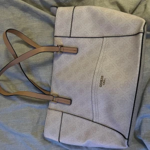 Beige axelväska från Guess - Snygg beige axelväska från Guess med logotypdetaljer och stilrena svarta kanter. Väskan har långa handtag i matchande färg och en praktisk dragkedja på insidan. Perfekt för att bära med sig allt man behöver under dagen. I nyskick och tror ej att den säljs längre. Tar emot nya bud