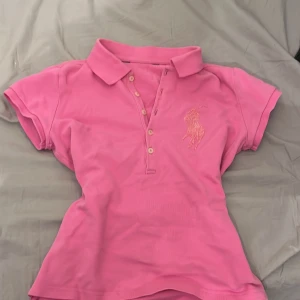 Rosa T-shirt från Ralph Lauren - Säljer en snygg rosa pikétröja från Ralph Lauren med klassisk krage och knappar framtill. Tröjan har korta ärmar och en broderad logga på bröstet. Perfekt för en avslappnad och stilren look.