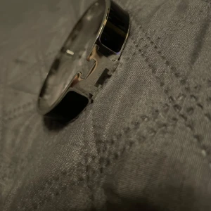 Silverfärgat armband med svart detalj - Snyggt och stilrent armband i silverfärg med en svart detalj. Armbandet har en unik design med ett H-format spänne som ger en modern touch. Perfekt för att komplettera din outfit med en elegant accessoar.