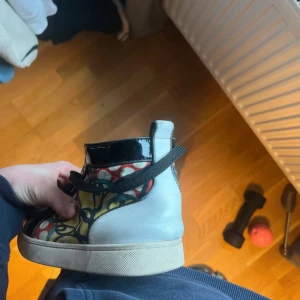 Färgglada sneakers med röd sula - Unika sneakers med färgglatt mönster och ikonisk röd sula. Skorna har snörning och en kombination av olika färger som svart, blå, gul och röd. Perfekta för den som vill sticka ut med sin stil.