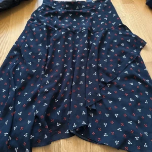 Blommig kjol i mörkblått - Charmig mörkblå kjol med små blommor i rött och vitt. Kjolen har en omlott-design som ger en fin rörelse och fall. Perfekt för en avslappnad stil med en touch av elegans.