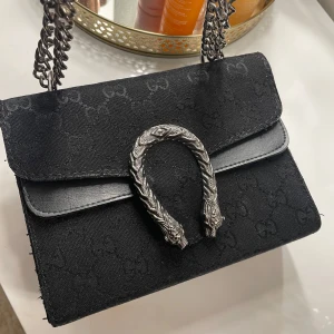 Svart axelväska med kedja - Snygg svart axelväska med en unik metallspänne framtill. Väskan har en kedjerem och ett diskret mönster i tyget. Perfekt för att ge en stilren touch till din outfit. Väskan är inte äkta,den är köpt från Turkiet 