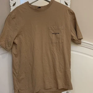 Beige t-shirt från H&M - Säljer en beige t-shirt från H&M med texten 'New York City U.S.A' på bröstfickan. T-shirten har en klassisk rund hals och korta ärmar. Perfekt för en avslappnad stil.