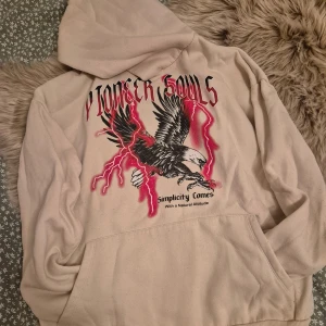 Beige hoodie med örntryck - Säljer en beige hoodie med ett coolt tryck av en örn och texten 'Pioneer Souls' i rött. Hoodien har en stor ficka framtill och en bekväm passform. Perfekt för en avslappnad stil.