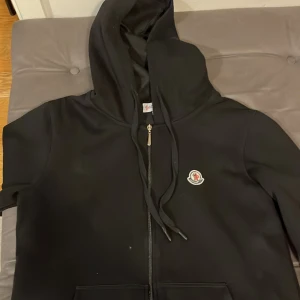 Svart hoodie från Moncler - Säljer en stilren svart hoodie från Moncler med dragkedja och justerbar huva. Den har en broderad logotyp på bröstet och är perfekt för en avslappnad look. Passar bra till både jeans och träningsbyxor.
