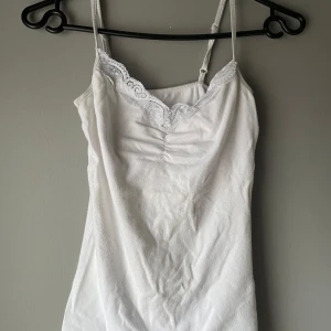 Vit linne med spetsdetaljer - Säljer ett stilrent vitt linne med smala axelband och vacker spetsdetalj vid halsringningen. Perfekt för varma dagar eller som ett lager under andra plagg.
