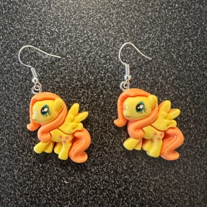 Färgglada örhängen med My little Pony  - Unika örhängen med en söt ponnydesign i gult och orange. Perfekta för att lägga till en lekfull touch till din stil. Ponnyerna har detaljerade ansikten och vingar, vilket gör dem till ett roligt och iögonfallande smycke.