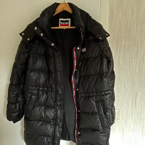Svart dunjacka från Levi's - Säljer en svart dunjacka från Levi's i storlek XS. Jackan har en pufferdesign med långa ärmar och en praktisk huva. Den har en dragkedja framtill och är perfekt för kyligare väder. Levi's-loggan syns på insidan och längs dragkedjan.