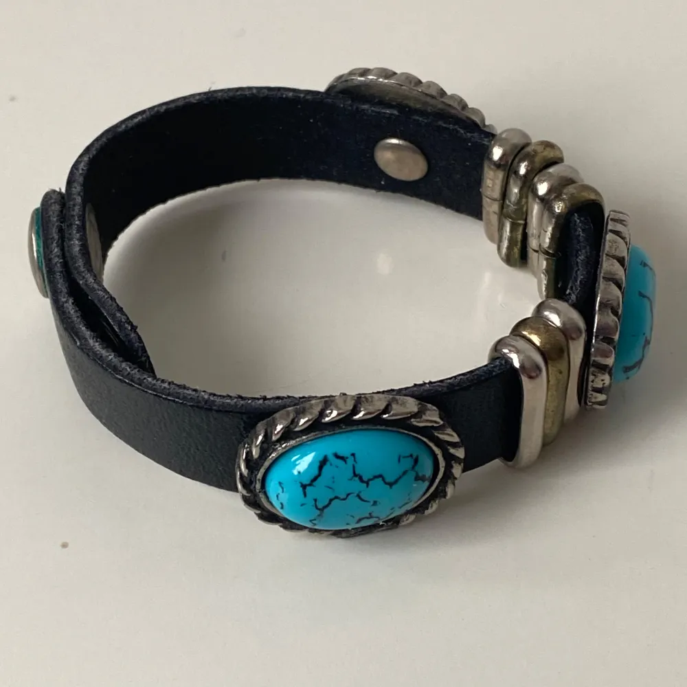 Snyggt svart armband med tre stora turkosa stenar infattade i silverfärgade detaljer. Ej äkta turkos. Armbandet har en justerbar tryckknappslåsning och metalliska dekorationer i guld och silver. Perfekt för att ge en bohemisk touch till din stil.. Asusteet.