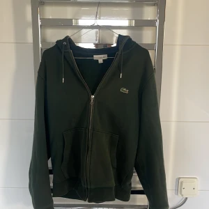 Grön hoodie från Lacoste - Säljer en snygg grön hoodie från Lacoste med dragkedja och huva. Den har det klassiska Lacoste-logotypen på bröstet och snören med märkets namn. Perfekt för en avslappnad stil.