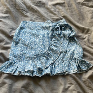 Blå blommig kjol från Shein - Söt blå blommig kjol från Shein med volangdetaljer och omlott-design. Kjolen har en dragkedja i sidan och en knytning framtill. Perfekt för en somrig look! 🌸Knytningen gör man själv 