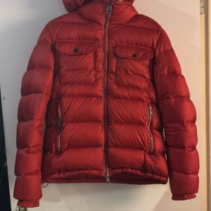 Röd Moncler Demar Giubbotto  - Säljer en snygg röd dunjacka från Moncler med huva och dragkedja. Huvan är avtagbar. Samt har Jackan två bröstfickor med knappar och två sidofickor med dragkedja. Perfekt för höst/vintern som nu börjar närma sig. Köpt från merchsweden