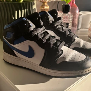 Nike Air Jordans i svart, blå och vit - Snygga Nike Air Jordans i en klassisk färgkombination av svart, blå och vit. Skorna har en ikonisk design med snörning och den välkända swoosh-loggan i blått på sidan. Perfekta för dig som vill ha en stilren och sportig look.