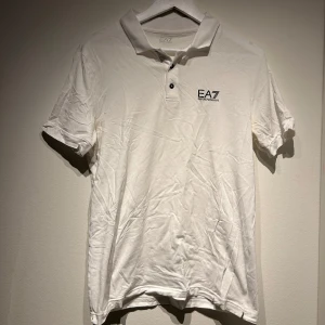 Vit pikétröja från EA7 Armani - Säljer en stilren vit pikétröja från EA7 Armani med korta ärmar och klassisk krage. Tröjan har en diskret logga på bröstet och knappar framtill. Perfekt för en avslappnad men ändå snygg look.