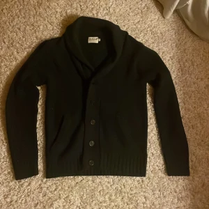 Svart kofta med knappar - Säljer en stilren svart kofta med knappar framtill. Den har en elegant krage och långa ärmar, perfekt för kyligare dagar. Koftan är ribbad vid ärmslut och nederkant vilket ger en snygg passform. Hör av dig vid frågor !