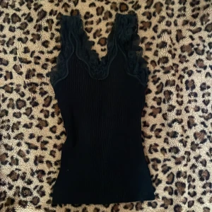 Svart ribbad linnetopp med spetsdetaljer - Snygg svart ribbad linnetopp med vacker spets runt ärmhålen och halsringningen. Skulle säga att det är i storlek S