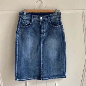 Blå jeanskjol med fickor - Snygg blå jeanskjol med klassisk femficksdesign och knappstängning fram. Kjolen är i mjuk och stretchig denim. Den har också dekorativa sömmar på bakfickorna. Midja 75 cm, längd 61. Står storlek L men är nog mer en M. Aldrig använd. 