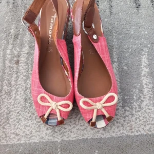 Rosa kilklackar från Tamaris - Snygga rosa kilklackar från Tamaris i bra skick. De har en söt rosett framtill och en bekväm kilklack i flätat material. Perfekta för sommarens fester eller en dag på stan. De har en öppen tå och justerbar rem runt hälen för bästa passform. Använda endast en gång!