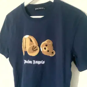 Säljer en mörkblå t-shirt från Palm Angels i storlek S. Den har ett coolt broderat nallemotiv på framsidan och texten 'Palm Angels' under. Perfekt för en avslappnad stil med en touch av lyx. T-shirten är i mycket bra skick och passar perfekt till både vardag och fest.