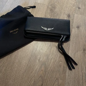 Svart väska från Zadig & Voltaire - Snygg svart väska från Zadig & Voltaire med en cool silvervinge-detalj på framsidan. Den har en dragkedja och kommer med en praktisk dustbag. Perfekt för att hålla ordning på dina kort och kontanter med stil! Världens finaste väska, inköpt tidigare i vår på tradera. Lång axelrem medföljer