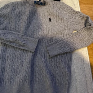 Helt Ny Grå kabelstickad tröja från Polo Ralph Lauren  - Säljer en snygg grå kabelstickad tröja från Polo Ralph Lauren. Den har en klassisk design med rund hals och det ikoniska logotypbroderiet på bröstet. Perfekt för både vardag och lite finare tillfällen. Tröjan är i nytt skick med etikett kvar. Passar perfekt till höst och vinter! Passar som xs med
