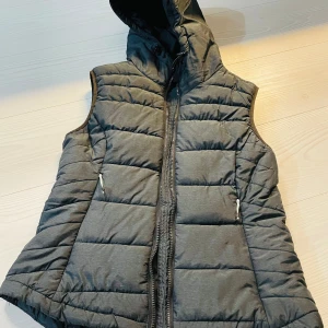 Grå dunväst från H&M Sport - Säljer en snygg grå dunväst från H&M Sport i storlek M. Västen har en praktisk dragkedja och en mysig huva, perfekt för kyliga dagar. Den är i mycket bra skick och passar utmärkt för höst och vår. Perfekt för en sportig look eller en avslappnad dag ute. Funkar lika bra till löprundan som en promenad inne i gallerian!