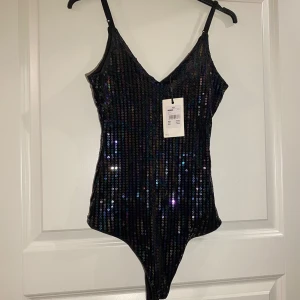 Svart glittrig body - Snygg svart body med glittriga paljetter, perfekt för festliga tillfällen. Den har smala axelband och en djup v-ringning som ger en elegant look. Bodyn är i nytt skick med taggen kvar. Perfekt för att glänsa på nästa fest! ✨
