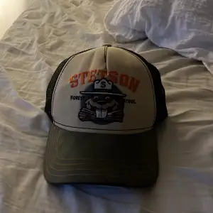 Säljer en cool Stetson keps med ett skogspatrullmotiv. Kepsen har en beige framsida med ett tryck av en bäver och texten 'Forest Patrol'. Baksidan är i svart mesh för bra ventilation. Perfekt för sommarens äventyr!