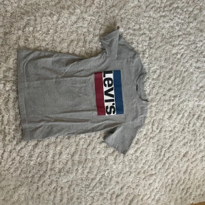 Grå t-shirt från Levi's - Säljer en grå t-shirt från Levi's i storlek 164 cm. Den har en klassisk design med en stor logga i rött, vitt och blått på framsidan. Perfekt för en avslappnad stil och passar till de flesta tillfällen. T-shirten är i bra skick och gjord av mjuk bomull.