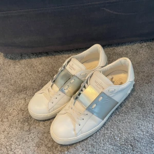 Valentino sneakers i vitt och silver - Säljer ett par snygga Valentino sneakers i vitt med en cool silverdetalj över snörningen. De är i mycket bra skick och perfekta för både vardag och fest. Skorna har en klassisk design med en modern twist tack vare den metalliska remmen.