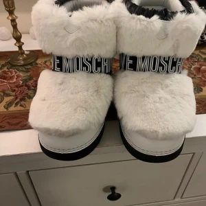 Vita pälsiga boots från Love Moschino - Superfina vita boots från Love Moschino med fluffig päls och svart textband runt skaftet. De har en cool design med ett rött hjärta på baksidan och är perfekta för vintern. Insidan är fodrad för extra värme och komfort. Perfekta för att hålla stilen även när det är kallt ute!