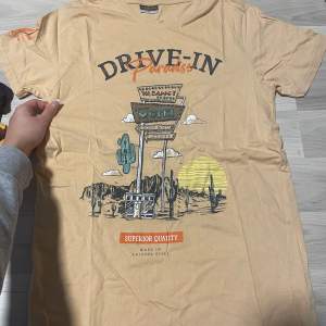 Säljer en beige t-shirt från Alphar One i storlek M. Den har ett coolt tryck med texten 'Drive-In Paradise' och en retro motelldesign. Perfekt för en avslappnad stil. T-shirten är kortärmad och gjord i ett mjukt material.