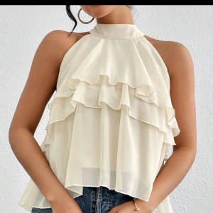 Beige ärmlös blus med volanger - Säljer en superfin beige blus med volanger. Den är ärmlös och har en luftig passform, perfekt för sommardagar. Materialet är lätt och skönt, vilket gör den idealisk för både vardag och fest. Passar perfekt med jeans eller kjol!
