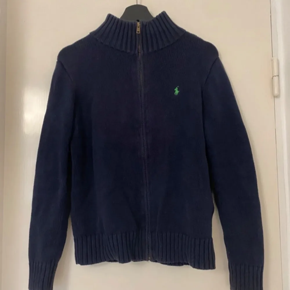 Säljer en snygg mörkblå stickad tröja från Polo Ralph Lauren. Den har en dragkedja framtill och en liten grön logga på bröstet. Perfekt för kyliga dagar och passar både till vardags och lite finare tillfällen. Tröjan har långa ärmar och en hög krage för extra värme.. Neuletakit & Villapaidat.