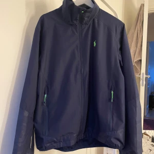Mörkblå jacka från Polo Ralph Lauren - Säljer en snygg mörkblå jacka från Polo Ralph Lauren. Den har en dragkedja framtill och två fickor med gröna detaljer. Jackan är perfekt för höst och vår, och har en stilren design med det klassiska Polo-loggan på bröstet. Passar både till vardags och lite finare tillfällen. 