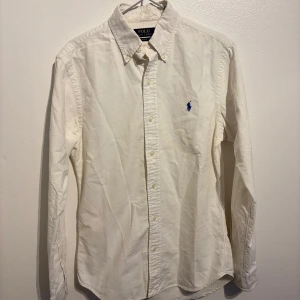  Ralph Lauren skjorta  - Säljer en klassisk vit skjorta från Ralph Lauren i slim fit. Skjortan har långa ärmar och är tillverkad i bomull. Den har en broderad blå logga på bröstet och knappar framtill. Perfekt för både vardag och fest! 🕺