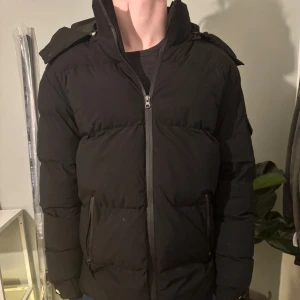 Svart pufferjacka med huva - Säljer en svart pufferjacka i mycket bra skick förutom på ett ställe där jag har lagat den syns inte vid användning då det är i under armen.Den har en dragkedja framtill och praktiska fickor. Jackan är perfekt för kalla dagar och har en skön huva. Passar  vardags. Perfekt för vintern!