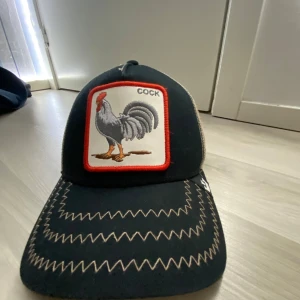 Svart och beige keps med tuppmotiv - Snygg svart keps med beige mesh-bakdel och ett broderat tuppmotiv framtill. Perfekt för sommardagar med sin luftiga design. Justerbar passform för att sitta bekvämt hela dagen.