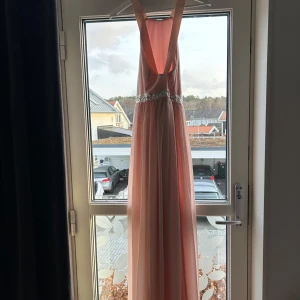 Rosa långklänning med strassdetaljer - Säljer en elegant rosa långklänning med vackra strassdetaljer i midjan. Klänningen har en öppen rygg och är axelbandslös, perfekt för festliga tillfällen. Materialet är mjukt och faller fint, vilket ger en lyxig känsla. Passar perfekt till bal eller nyårsfest! 🌟