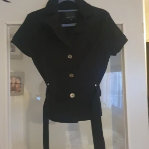 Svart kortärmad kavaj med bälte - Snygg svart kavaj med korta ärmar och knappar framtill, mjukare tyg. Den har ett bälte i midjan för en fin passform. Perfekt för både vardag och fest!