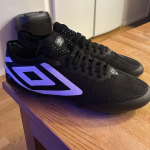 Svarta fotbollsskor från Umbro - Säljer ett par svarta fotbollsskor från Umbro i nytt skick. Skorna har en snygg design med vita detaljer och snörning. De är perfekta för fotbollsträning och matcher. Sulan har flera dobbar för bra grepp på planen. Passar dig som vill ha stil och funktion i ett. Skorna är helt splitter nya