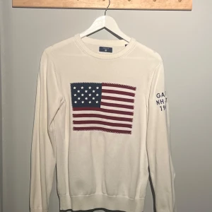 Vit stickad tröja med amerikansk flagga från GANT - Säljer en stilren vit tröja från GANT i storlek S. Den har en stor amerikansk flagga på framsidan och text på ärmen, där det står gant nh-ct 1949. Perfekt för en casual look! Tröjan är i mycket bra skick och passar alla årstider och tillfällen. Öppen för byten! Priset kan diskuteras vid snabb affär.