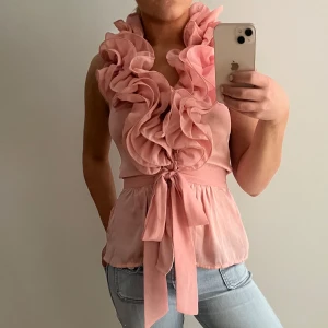 Rosa blus med volangdetaljer - Säljer en superfin rosa blus med stora volangdetaljer framtill och en knytning i midjan. Den sitter runt nacken och är alltså ärmlös