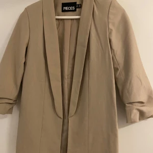 Beige kavaj från Pieces - Snygg beige kavaj från Pieces i ett stilrent snitt. Perfekt för både vardag och fest. Den har trekvartsärmar och en klassisk krage. Passar bra till både jeans och klänning för en chic look.