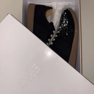 Svarta sneakers med färgstänk Från Maison Margiela - Säljer ett par svarta sneakers med coola färgstänk i vitt, blått och gult. Skorna har en klassisk design med snörning och en bekväm brun sula. Perfekta för att ge din outfit en unik touch! De är i nytt skick och kommer i originalförpackning. PERFEKTA JULKLAPPEN FÖR TÅNÅRINGAR