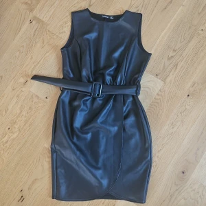Svart ärmlös klänning  med bälte - Snygg svart ärmlös klänning i skinnimitation från Boohoo. Den har en tight passform och ett matchande bälte i midjan för en stilren look. Strechig i materialet.  Perfekt för en utekväll eller fest! 🖤